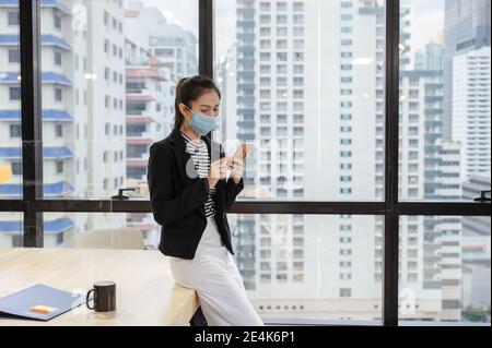 Young asian woman manager wearing face mask using smartphone during lunch break in modern office on downtown Stock Photo