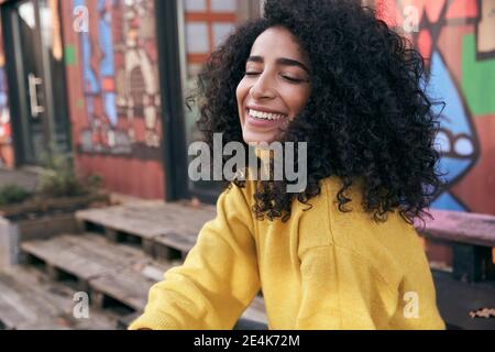 Smiling young woman with eyes closed sitting against wall in city Stock Photo