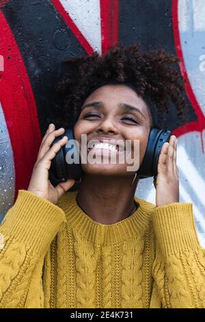 Happy young woman with eyes closed listening music through headphones against painted wall