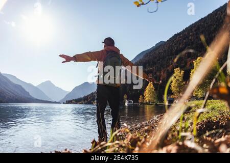 Mature male hiker with arms outstretched looking at lake against clear sky during sunny day Stock Photo