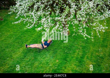 Man reading book sitting on chair under tree in yard Stock Photo