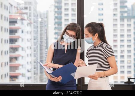 Multi ethnic two young business colleague wearing face mask discussing about business in modern office at side window Stock Photo