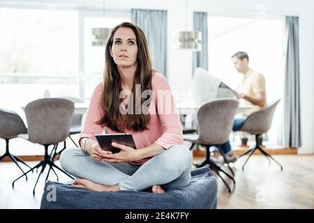 Mature woman with man in background day dreaming while holding digital tablet at home Stock Photo