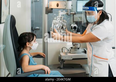 Mid adult female optometrist adjusting propther for medical test at clinic during COVID-19 Stock Photo
