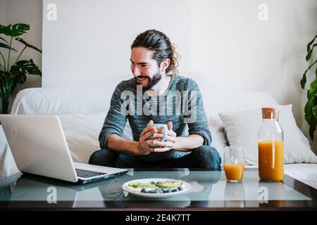 Young man with coffee cup using laptop while having breakfast sitting on sofa at home Stock Photo