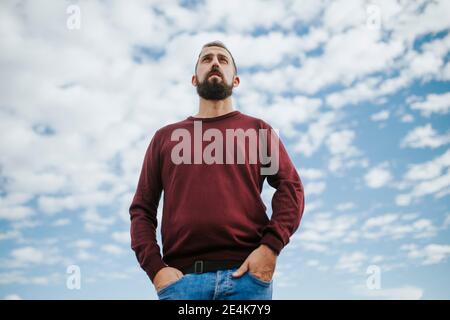 Thoughtful bearded man with hands in pockets standing against cloudy sky Stock Photo
