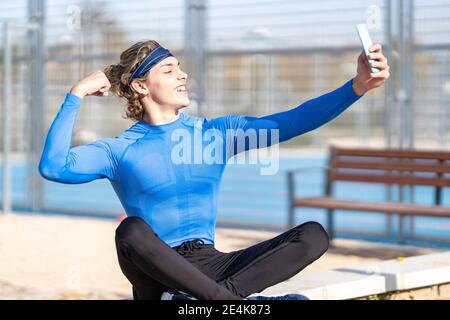 Smiling male athlete showing bicep while taking selfie i public park on sunny day Stock Photo