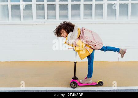 Portrait of cheerful girl with curly hair riding push scooter on road by building Stock Photo