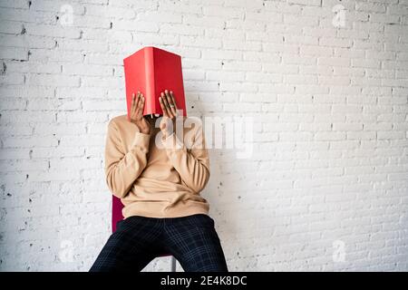 Man covering face with red book while sitting on chair against white brick wall Stock Photo