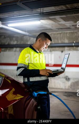 Smiling male technician with laptop using mobile phone by electric car at auto repair shop Stock Photo
