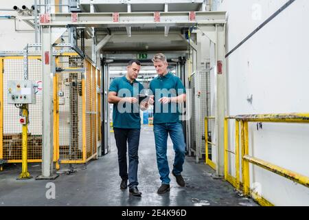 Mature male engineer colleagues walking while discussing over digital tablet in factory Stock Photo