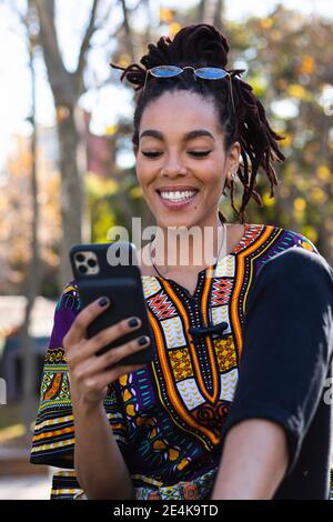 Happy fashionable young woman using mobile phone while sitting at park Stock Photo