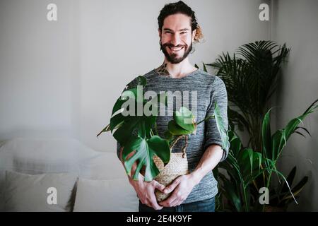 Young man smiling while holding Monstera plant at home Stock Photo