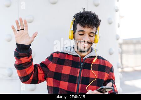 Smiling man wearing headphones waving hand while using mobile phone standing against wall Stock Photo