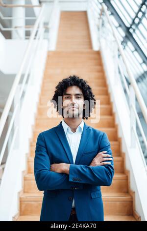 Confident handsome young male entrepreneur standing with arms crossed against staircase at office Stock Photo