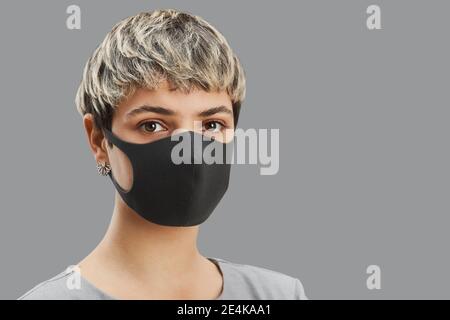 Head and shoulders portrait of contemporary short-haired woman wearing mask while standing against minimal gray background, copy space Stock Photo