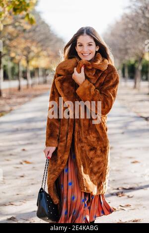 Happy woman in fur coat walking while holding purse on footpath Stock Photo