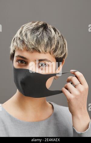 Vertical head and shoulders portrait of contemporary short-haired woman taking off mask while standing against minimal gray background, copy space Stock Photo