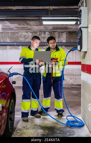 Male programmer discussing with colleague over laptop while charging electric car Stock Photo