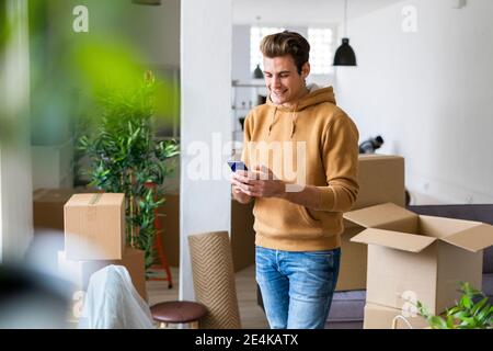 Smiling young man using mobile phone in messy living room during relocation Stock Photo