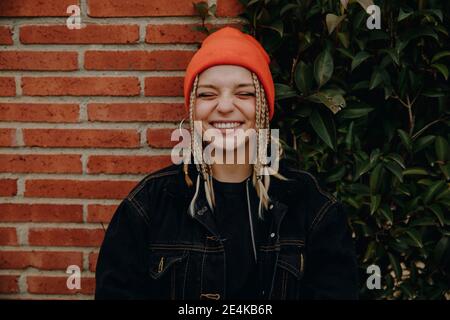 Cheerful young woman with eyes closed standing against plants and brick wall Stock Photo