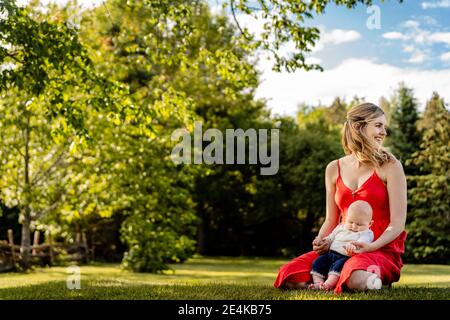 Smiling mother holding baby boy while kneeling on grassy land in park Stock Photo