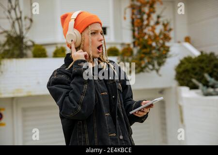 Young woman with smart phone giving shocking expression while listening music in front yard Stock Photo