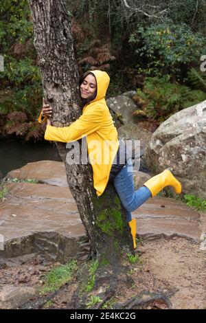 Happy female hiker wearing yellow raincoat embracing tree in forest Stock Photo