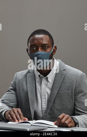 Vertical portrait of confident African-American businessman wearing mask and looking at camera while sitting at desk against minimal gray background, Stock Photo