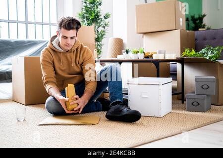 Young man wrapping drinking glass in brown paper while moving in new apartment Stock Photo