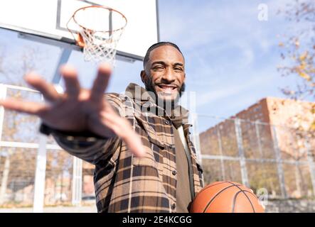 Happy young man gesturing while holding basketball in court on sunny day Stock Photo