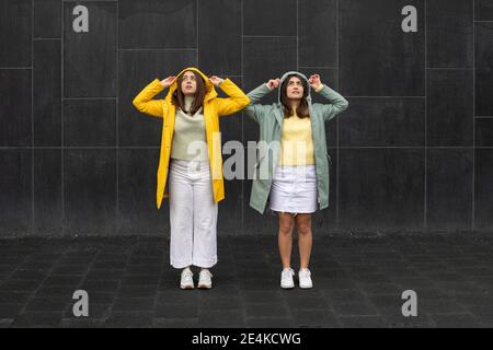 Young women wearing raincoats looking up while standing against black wall Stock Photo