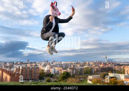 Male professional wearing pig mask taking selfie from smart phone while jumping in city against sky Stock Photo