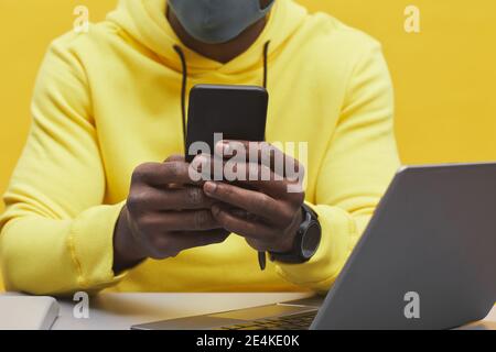 Cropped shot of unrecognizable African-American man holding smartphone while sitting at desk against pop yellow background Stock Photo