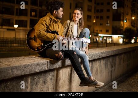 Couple with guitar smiling while sitting on retaining wall Stock Photo