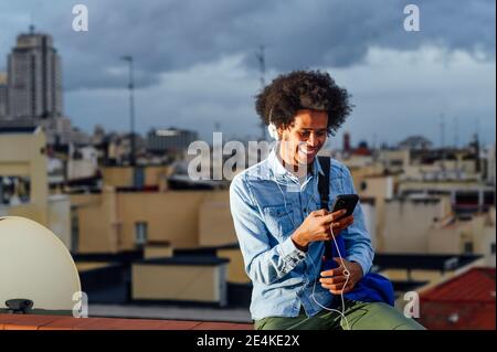 Happy young man with curly hair using smart phone against buildings in city Stock Photo