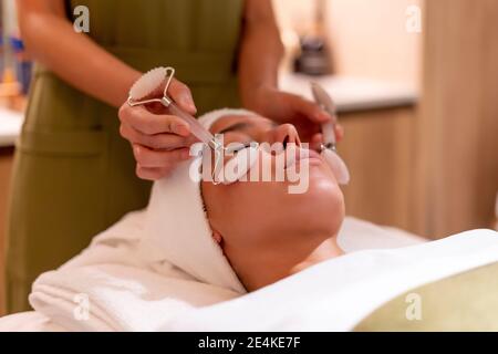 Beautician holding gemstone rollers on young female customer's face during treatment at health spa Stock Photo