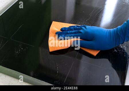 men's hand in blue glove washes the modern electric hob with a sponge . Close-up view Stock Photo