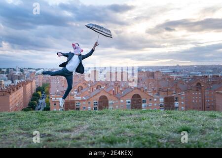 Male professional wearing pig mask holding umbrella while jumping on hill at sunset Stock Photo