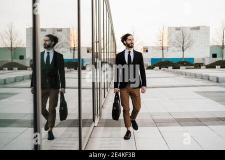 Young businessman with briefcase looking away while walking by glass wall Stock Photo
