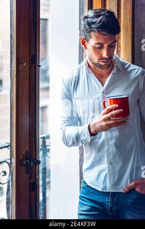 Thoughtful young man holding cup standing at the window Stock Photo