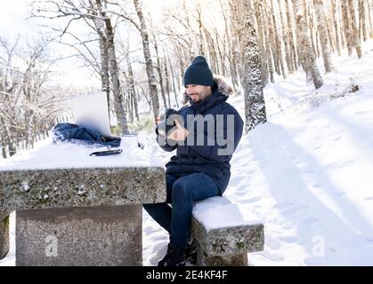 Smiling photographer using camera while sitting on rock bench in forest during snowy weather Stock Photo