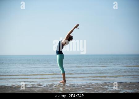 Mature woman with arms raised exercising at beach against clear sky Stock Photo