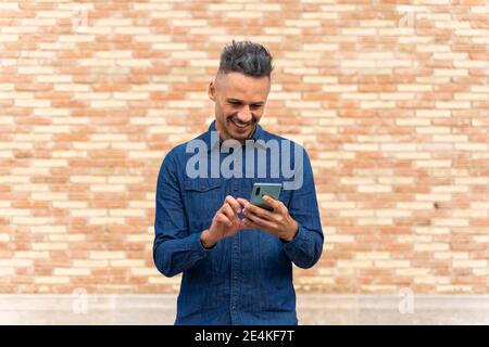 Happy man using smart phone against brick wall Stock Photo