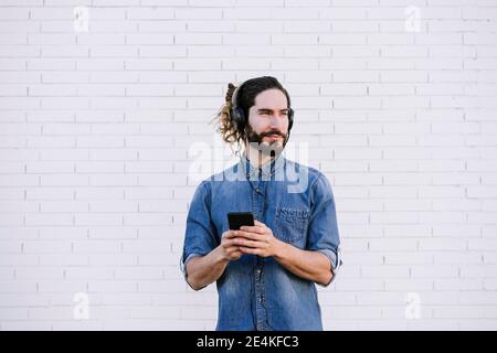 Stylish young man listening music through headphones while looking away against brick wall Stock Photo