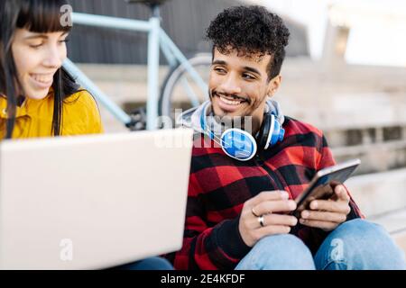 Smiling friends using laptop and digital tablet while sitting outdoors Stock Photo
