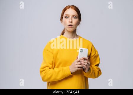 Portrait of confused young woman wearing yellow sweater holding mobile phone and looking at camera Stock Photo