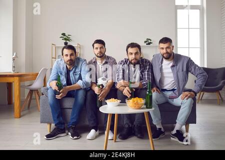 Group of friends sitting on sofa with chips and beer and watching soccer game on TV Stock Photo