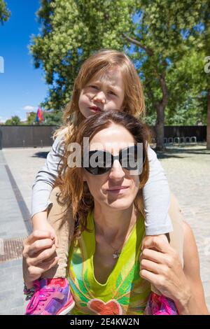portrait of woman mother with black sunglasses carrying on shoulders a three years old blonde child, tired or lazy, on urban street of Madrid city, Sp