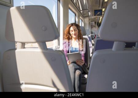 red hair happy woman dressed in purple and blue, traveling by train sitting reading digital tablet or ebook Stock Photo